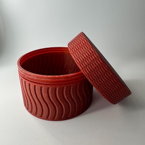 May include: A red cylindrical container with a textured lid. The container has a wavy pattern and the lid has a rope-like pattern.