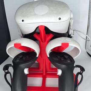 Op de afbeelding: Een witte VR-headset en controllers worden getoond op een rode standaard. De controllers zijn zwart met witte accenten. De standaard is ontworpen om de headset en controllers te bewaren.
