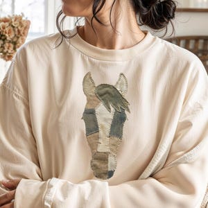 May include: Cream-colored sweatshirt featuring a horse head design. The horse head is made of various fabric patches in shades of beige, gray, and green. The sweatshirt has a crew neck and long sleeves with ribbed cuffs.