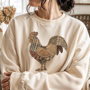 May include: Cream-colored sweatshirt featuring a patchwork rooster design. The rooster is made of various fabric patterns, including plaid and floral prints. The sweatshirt has a crew neck and long sleeves. The overall style is rustic and country-inspired.
