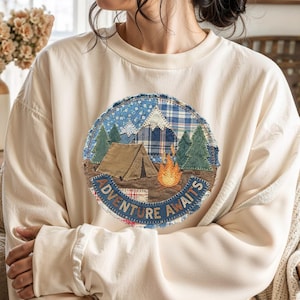 May include: Cream-colored sweatshirt with a circular patch featuring a camping scene. The patch includes a tent, campfire, trees, and the words "Adventure Awaits." The design incorporates blue plaid and floral patterns. The sweatshirt has long sleeves.