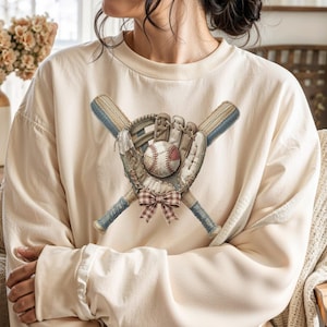Peut inclure: Sweat-shirt crème avec un motif de baseball. L'image présente un gant de baseball, une balle et des battes croisées. Les battes sont bleu clair avec des manches marron, et un nœud à carreaux est noué en dessous. Le sweat-shirt a une coupe décontractée.