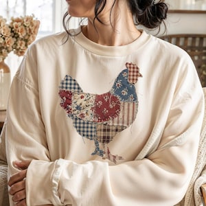 Könnte beinhalten: Cremefarbenes Sweatshirt mit einem Patchwork-Hühnerdesign aus roten, blauen und braunen Stoffen. Das langärmlige Oberteil hat einen Rundhalsausschnitt und gerippte Bündchen. Das Hühnerdesign ist mittig auf der Vorderseite des Shirts platziert.