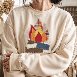 May include: Cream-colored sweatshirt featuring a colorful campfire applique. The applique includes red, orange, yellow, and plaid fabric flames, with brown, blue, and white log details. The sweatshirt has long sleeves and a relaxed fit.