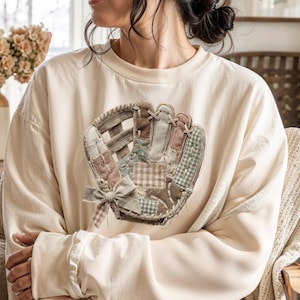 Könnte beinhalten: Cremefarbenes Sweatshirt mit einem Baseballhandschuh-Design. Der Handschuh besteht aus Patchwork-Stoff in Braun-, Rosa- und Blautönen mit einer Schleife. Das Sweatshirt hat lange Ärmel und eine lockere Passform, ideal für Freizeitkleidung.