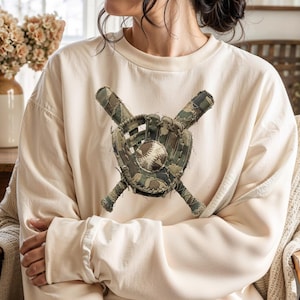 May include: Cream-colored sweatshirt featuring a baseball glove and crossed bats design. The baseball glove and bats are made of a camouflage pattern. The sweatshirt has long sleeves and a crew neck.