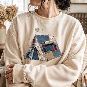 Może przedstawiać: Kremowa bluza z aplikacją w kształcie namiotu wykonanego z patchworku. Namiot wykonany jest z różnych tkanin, w tym dżinsu i kwiatowych wzorów, z małą flagą na górze. Bluza ma okrągły dekolt i długie rękawy.