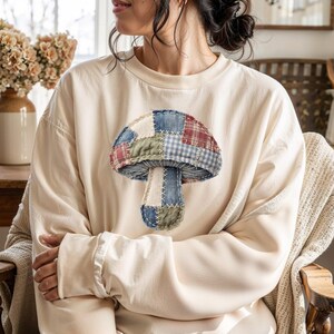 May include: Cream-colored sweatshirt featuring a patchwork mushroom design. The mushroom cap is composed of various fabric patterns in red, blue, green, and white. The stem is a light beige color. The sweatshirt has long sleeves and a relaxed fit.