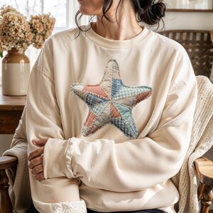 May include: Cream-colored sweatshirt featuring a large, patchwork star design in shades of pink, blue, and green. The star is made of various fabric patterns, including gingham and solid colors. The sweatshirt has a relaxed fit and long sleeves.