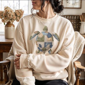 Op de afbeelding: Crèmekleurige sweatshirt met een patchwork schildpadontwerp in blauw, groen en bruin. De schildpad is gecentreerd op de voorkant van de top met lange mouwen. De sweatshirt heeft een relaxte pasvorm en geribde manchetten.