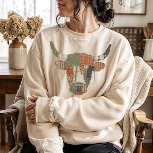 May include: Cream-colored sweatshirt featuring a patchwork bull head design. The bull's head is composed of various fabric patches in shades of green, brown, and black. The sweatshirt has long sleeves and a crew neck. The overall style is casual and rustic.