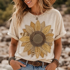Puede incluir: Camiseta de color crema con un gran diseño de girasol en la parte delantera. El girasol está hecho de varios parches de tela en tonos amarillos, marrones y verdes. La camiseta tiene mangas cortas y cuello redondo.