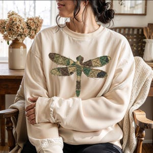 May include: Cream-colored sweatshirt featuring a large dragonfly design. The dragonfly is made of patchwork fabric in shades of green, brown, and tan. The sweatshirt has long sleeves and a relaxed fit, suitable for casual wear.