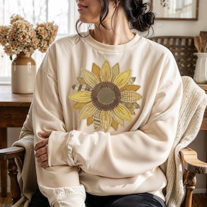 May include: Cream-colored sweatshirt featuring a large sunflower applique. The sunflower has layered petals in shades of yellow and brown, with a textured center. The sweatshirt has long sleeves and a relaxed fit, perfect for casual wear.