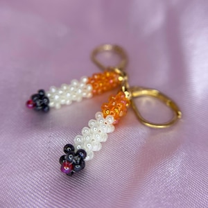Op de afbeelding: Een paar kralen oorbellen in de vorm van sigaretten. De oorbellen hebben witte, oranje en zwarte kralen, met goudkleurige oorhaken. De achtergrond is roze.