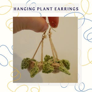 May include: A pair of crocheted hanging plant earrings. Each earring features a small, beige pot with green crocheted leaves. The earrings are suspended from gold hooks.
