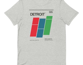 May include: A grey t-shirt with a retro VHS tape design featuring the word "DETROIT" in white text and a colorful graphic of three bars in red, blue, and green. The text "VHS" is in white on a black rectangle in the upper right corner. The text "VIDEO CASSETTE" is in black below the graphic.