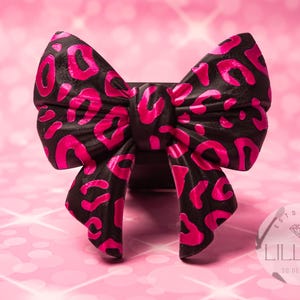 May include: A decorative bow featuring a black base with a vibrant hot pink leopard print pattern. The bow has a structured, folded design with two main loops and two downward-hanging tails. A circular logo with the text LILLIAN 3D DESIGNS is visible.