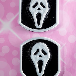 May include: Two white and black 3D printed Ghostface masks from the Scream movie franchise. The masks are on a pink background.