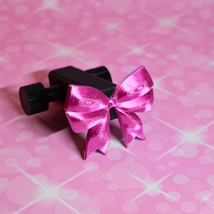 May include: A black plastic clamp with a bright pink bow attached to it. The clamp is designed to hold objects in place.