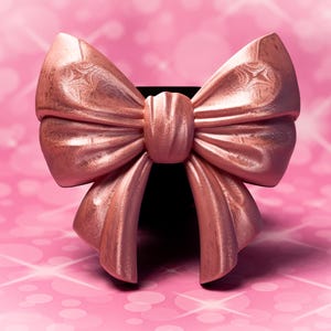 May include: A large, rose gold bow with detailed textures. The bow is set against a soft pink background with a bokeh effect. The bow's shape and finish suggest it is made of a luxurious material, suitable for gift wrapping or decorative use.
