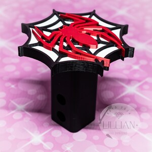 May include: Black and red 3D printed hitch cover featuring a spider in a web. The hitch cover has the text "ESTD 2024 LILLIAN+ 3D DESIGNS" on the bottom.