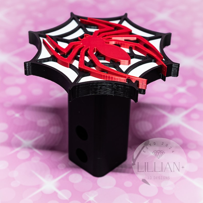 May include: Black and red 3D printed hitch cover featuring a spider in a web. The hitch cover has the text "ESTD 2024 LILLIAN+ 3D DESIGNS" on the bottom.