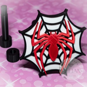 May include: A 3D printed red spider on a white web with a black border. The spider is in the center of the web and the web is in the shape of a circle. The web is on a pink background.