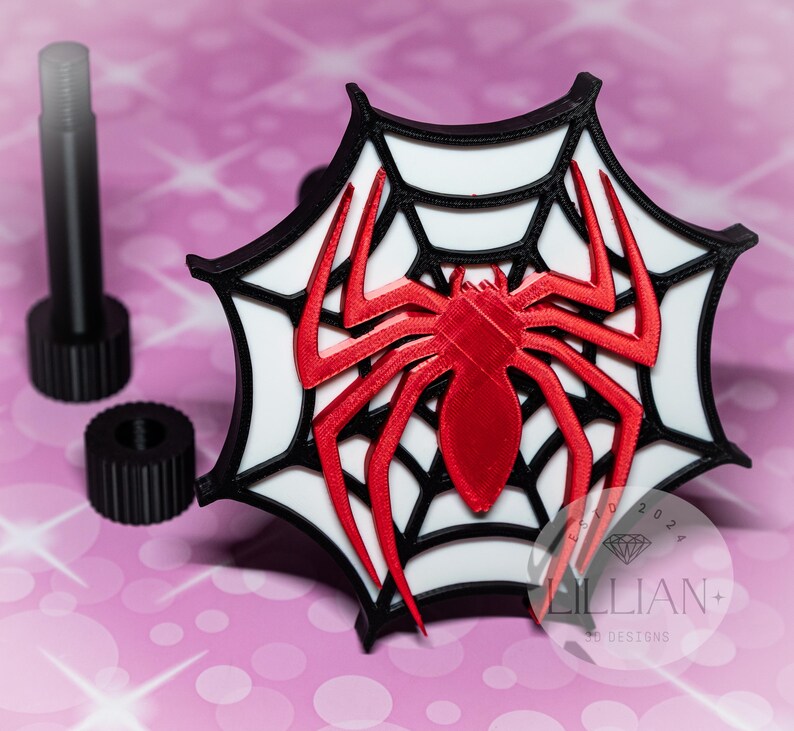 May include: A 3D printed red spider on a white web with a black border. The spider is in the center of the web and the web is in the shape of a circle. The web is on a pink background.