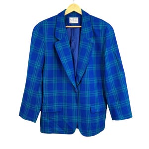 Puede incluir: Un blazer a cuadros azul y verde azulado con cierre de un solo botón y dos bolsillos delanteros. El blazer está colgado de una percha de madera. El forro interior es de color azul liso. La etiqueta es visible.