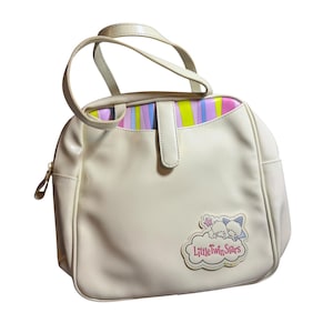 Könnte beinhalten: Cremefarbene Handtasche mit oberem Griff und Klappenverschluss. Der Innenraum hat ein Streifenmuster in Rosa, Lila, Grün und Blau. Ein Aufnäher auf der Vorderseite zeigt den Text "Little Twin Stars" mit einer Wolken- und Sterngrafik.