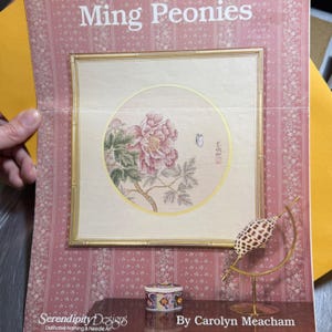 Puede incluir: Una obra de arte enmarcada titulada "Ming Peonies" con una flor de peonía rosa dentro de un círculo enmarcado en oro. La obra de arte está sobre un fondo rosa con estampado floral. También se ve el texto "Serendipity Designs" y "By Carolyn Meacham".
