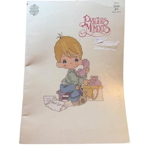 May include: A cross-stitch pattern featuring a blonde-haired child with a sad expression, holding a pink telephone. The design includes the text "Precious Moments" and "Dear Jon". The background is a light beige color.