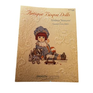 May include: A book cover for "Antique Bisque Dolls" with a cross-stitch illustration of a doll, dog, and toy. The design uses blue, red, and brown. The cover has a decorative lace border.