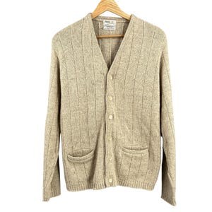 May include: A beige, button-down cardigan sweater with a V-neck and two front pockets. The sweater is hanging on a wooden hanger. The sweater has a ribbed design.