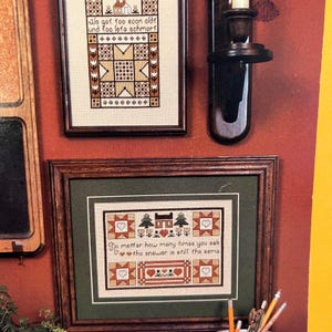 May include: Two framed cross-stitch samplers with brown wooden frames are displayed on a red wall. The top sampler has text and geometric patterns. The bottom sampler features text and heart, tree, and house motifs.