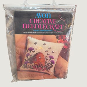 Könnte beinhalten: Ein Avon Creative Needlecraft Crewel-Stickset mit dem Titel The Bee Garden. Die Verpackung zeigt ein beiges Kissen mit einem Bienenstock, Bienen und violetten Blumen. Der Text besagt, dass alles außer einem 41 cm mal 41 cm Kissen enthalten ist.