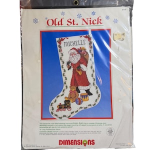 May include: A cross-stitch kit with a Christmas stocking design. The design features Santa Claus, a teddy bear, a toy train, and the name "MICHELLE". The kit is packaged in a clear plastic bag and includes the text "Old St. Nick". Finished size is 40cm.