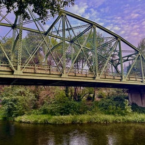 Puede incluir: Un puente de metal verde cruza un río, con vegetación exuberante a ambos lados. El puente tiene un diseño de celosía y está sostenido por pilares de hormigón.