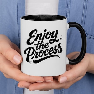 May include: White ceramic mug with a black interior and handle. The mug features the phrase "Enjoy the Process" in black cursive lettering, with star accents. The mug is held by a person.