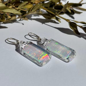 Puede incluir: Un par de pendientes rectangulares con un diseño transparente e iridiscente. Los pendientes tienen un cierre de palanca de color plateado. El efecto iridiscente crea un arcoíris de colores dentro del material transparente. Los pendientes se muestran sobre una superficie blanca.