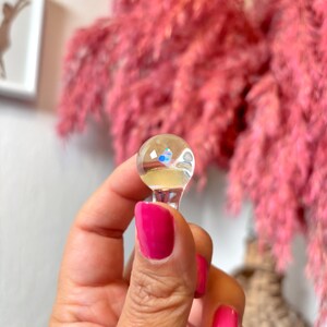 May include: A clear glass bottle stopper with a small, iridescent blue and white design inside. The stopper has a round top and a short, clear stem.