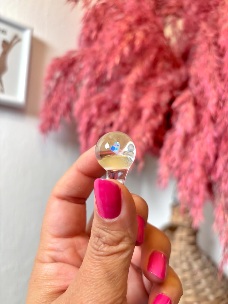 May include: A clear glass bottle stopper with a small, iridescent blue and white design inside. The stopper has a round top and a short, clear stem.