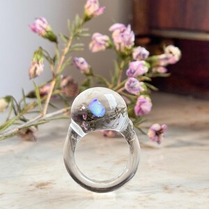 May include: A clear glass ring with a blue iridescent stone embedded in the center. The ring is sitting on a white surface with a sprig of pink flowers in the background.