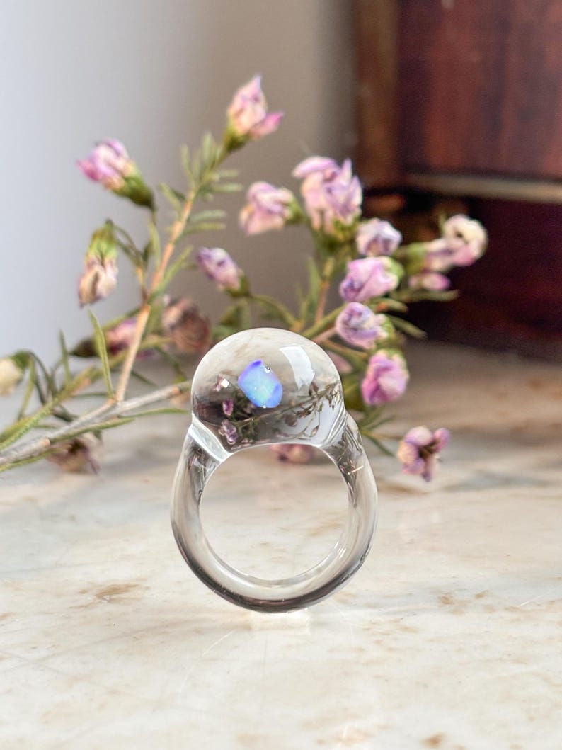 May include: A clear glass ring with a blue iridescent stone embedded in the center. The ring is sitting on a white surface with a sprig of pink flowers in the background.