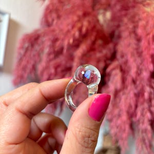 May include: Clear glass ring with a round, iridescent stone. The ring is thick and has a wide band.