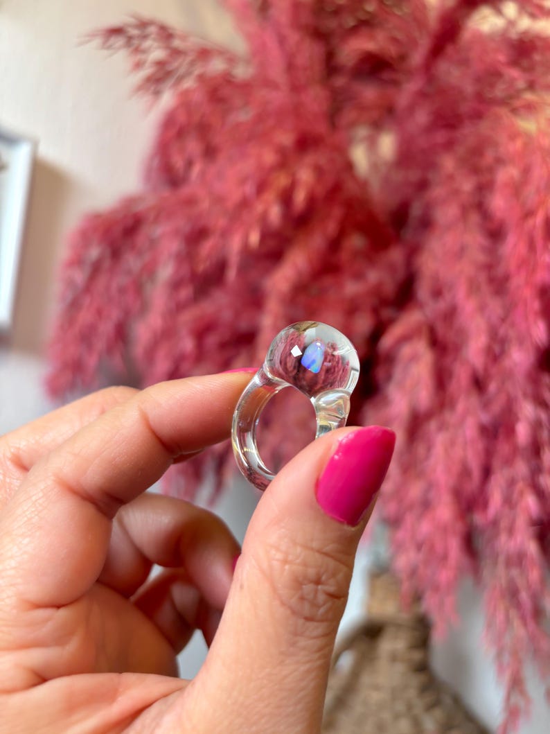 May include: Clear glass ring with a round, iridescent stone. The ring is thick and has a wide band.