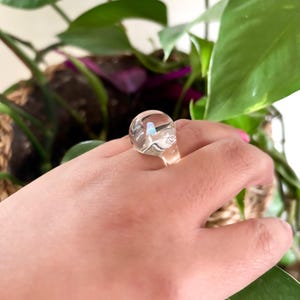 May include: A clear glass ring with a round, slightly iridescent stone. The ring is on a person's finger.