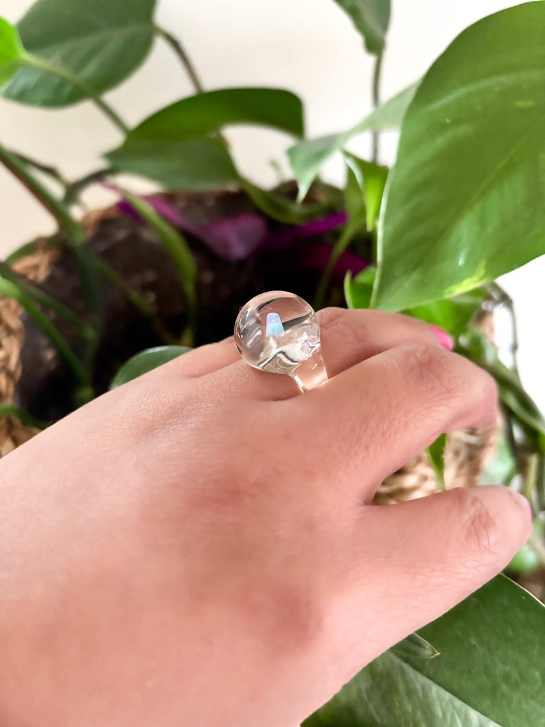 May include: A clear glass ring with a round, slightly iridescent stone. The ring is on a person's finger.