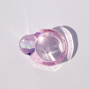 Könnte beinhalten: Ein durchscheinender, hellvioletter Ring mit einem abgerundeten, oval geformten Akzent. Das Ringband ist ein hellerer Lilaton, und der Akzent enthält schillernde Schimmer. Der Ring wirft einen Schatten auf eine weiße Oberfläche.